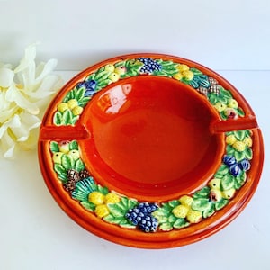 May include: A round ceramic ashtray with a glossy orange center and a raised rim decorated with a relief pattern of green leaves, yellow berries, and clusters of dark blue grapes. The design features two notches for resting items on the rim.