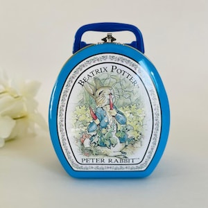 Puede incluir: Una fiambrera de metal azul con una ilustración de Peter Rabbit de Beatrix Potter. La caja ovalada tiene un asa azul y un cierre plateado. La ilustración muestra a Peter Rabbit sosteniendo zanahorias en un jardín.