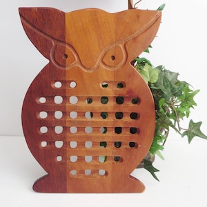 May include: A wooden owl-shaped trivet featuring a multi-toned brown finish with a natural wood grain. The design includes carved eye details at the top and a grid of square cutouts across the center body, creating a functional heat-resistant surface.
