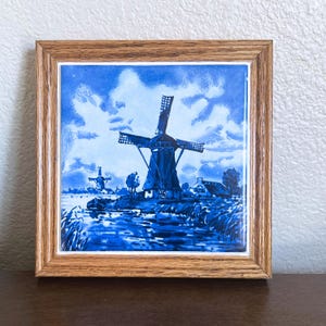 May include: A square ceramic tile featuring a blue and white illustration of a Dutch windmill landscape with a canal, trees, and a small house, encased in a light brown wooden frame with a visible wood grain texture and a simple beveled edge.
