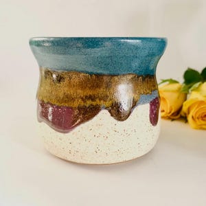 May include: A ceramic planter with a flared rim featuring a multi-coloured glaze. The top section is teal, transitioning into a band of metallic bronze, followed by a white base with dark speckles and drips of deep purple glaze throughout the surface.