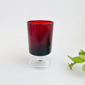 May include: A deep red glass goblet with a smooth, cylindrical bowl and a clear glass pedestal base. The vessel features a solid, dark crimson color throughout the upper portion, contrasting with the transparent, circular foot at the bottom.