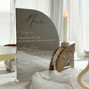 Könnte beinhalten: Eine reflektierende, halbkreisförmige Menütafel mit graviertem Text, darunter "Menu" und eine Liste von Gerichten. Rechts daneben eine Kerze, neben einer Tischnummer "2" in einem Steinständer. Das Menü ist auf Deutsch.