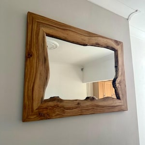 May include: A rectangular mirror with a wooden frame featuring a natural, live edge design. The frame is made of light brown wood, showing grain and knots. The mirror is mounted on a light grey wall.