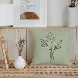 May include: A sage green square pillow with a black line drawing of leaves. The pillow is on a wooden bench. A framed botanical print and a white vase with dried flowers are on a shelf above.