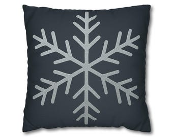 Puede incluir: Un cojín cuadrado con fondo gris oscuro. Un gran diseño de copo de nieve plateado está centrado en el cojín. El copo de nieve tiene seis brazos simétricos con puntas detalladas, creando un tema invernal.