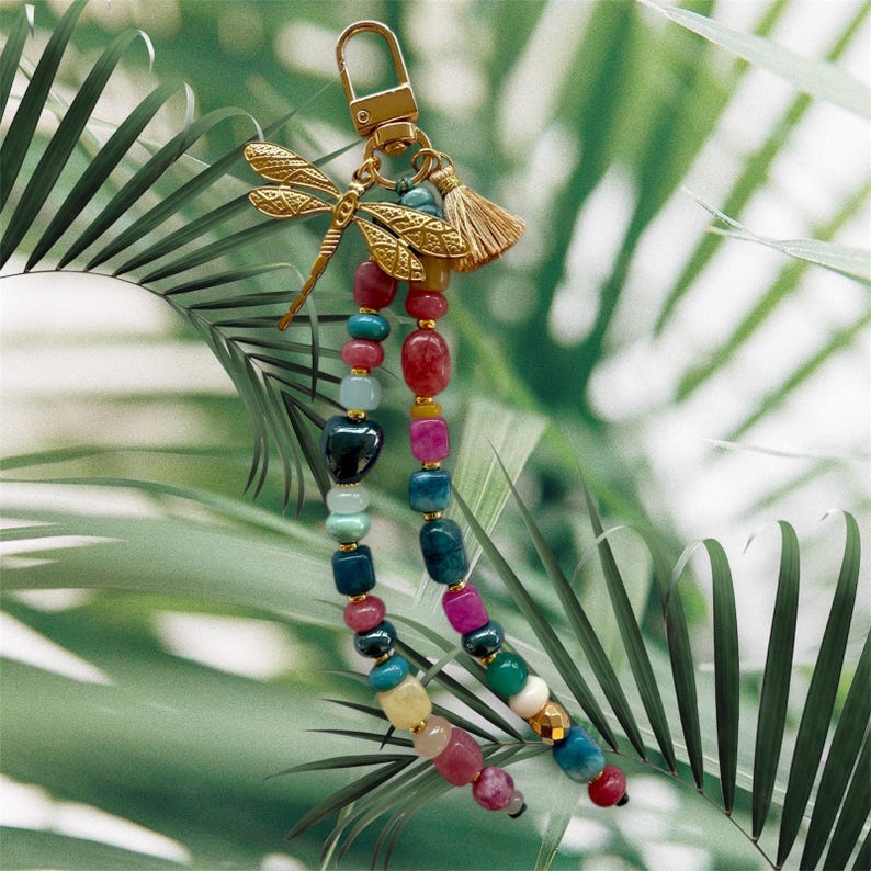 May include: A gold-colored keychain with a dragonfly charm and a small tassel. The keychain features two strands of colorful beads in various shapes and sizes, including red, blue, green, and pink.