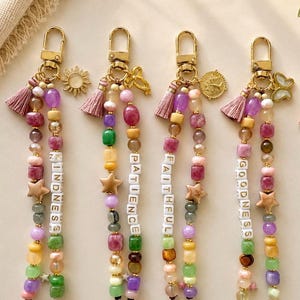 May include: Four beaded keychains with gold-tone lobster clasps and pink tassels. Each features multi-colored gemstone beads, a gold star charm, and white letter beads spelling KINDNESS, PATIENCE, FAITHFUL, and GOODNESS with unique charms.