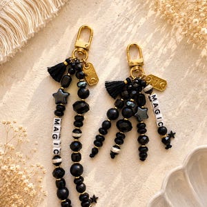 May include: Two gold-toned metal keychains featuring black beads, small black tassels, and rectangular charms with sun and moon engravings. One keychain has two long beaded strands, while the other has multiple shorter strands. Both include white letter beads spelling MAGIC.