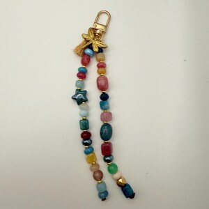 May include: A beaded keychain with a gold-colored clasp and a small tassel. The keychain features a variety of colorful beads, including blue, red, yellow, and green, and a dark blue star-shaped bead. A gold-colored dragonfly charm is attached.