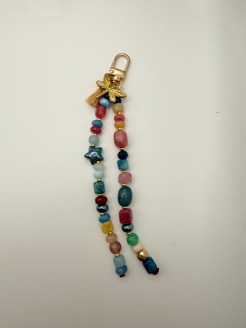 May include: A beaded keychain with a gold-colored clasp and a small tassel. The keychain features a variety of colorful beads, including blue, red, yellow, and green, and a dark blue star-shaped bead. A gold-colored dragonfly charm is attached.