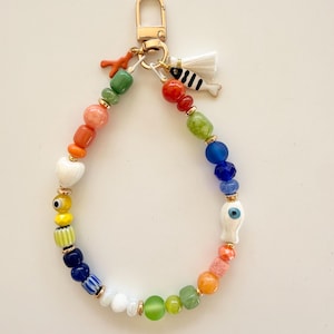 May include: A colorful beaded keychain with a gold clasp. The beads are in various shapes and colors, including red, orange, yellow, green, blue, and white. Charms include a coral, a fish, and a white tassel.