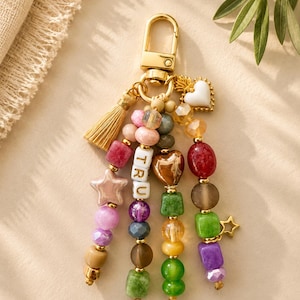 May include: A gold-toned metal keychain featuring a lobster clasp and a gold tassel. Four strands of multi-colored beads hang from the ring, including pink, purple, green, and brown shapes. One strand features white letter beads spelling TRU.