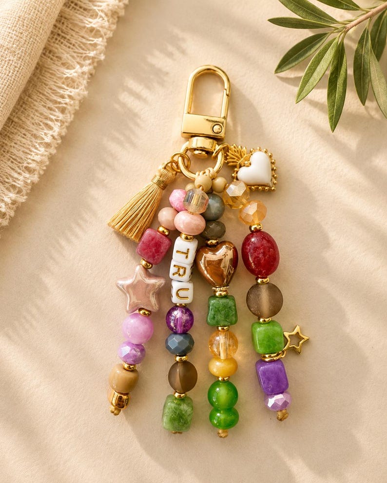 May include: A gold-toned metal keychain featuring a lobster clasp and a gold tassel. Four strands of multi-colored beads hang from the ring, including pink, purple, green, and brown shapes. One strand features white letter beads spelling TRU.