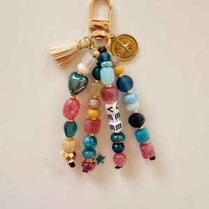 May include: A beaded keychain with a gold-colored clasp, tassel, and charm. The keychain features strands of colorful beads in shades of pink, blue, green, and gold. The word "LOVE" is spelled out in white letter beads.