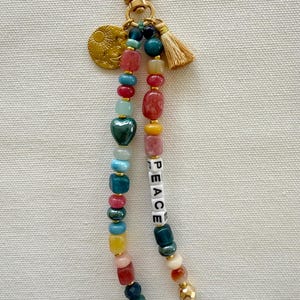 May include: A beaded keychain with a gold-tone clasp, a small tassel, and a charm with a sun and moon design. The keychain features colorful beads, including a green heart-shaped bead, and the word "PEACE" spelled out in white letter beads.