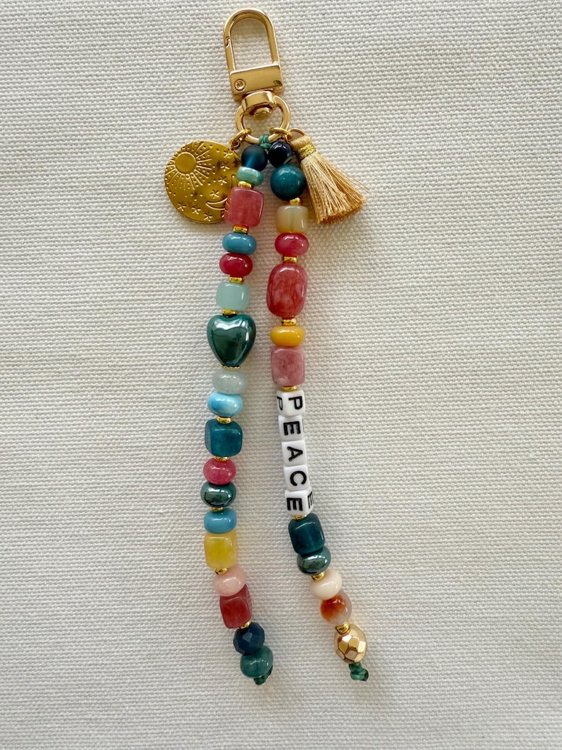 May include: A beaded keychain with a gold-tone clasp, a small tassel, and a charm with a sun and moon design. The keychain features colorful beads, including a green heart-shaped bead, and the word "PEACE" spelled out in white letter beads.