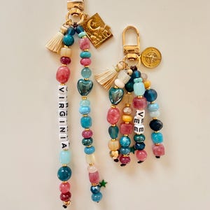 May include: Two beaded keychains with gold-tone hardware. One keychain spells "VIRGINIA" with white letter beads. The other keychain spells "VEE". Both keychains feature colorful beads, tassels, and charms, including a gold star and a crescent moon.