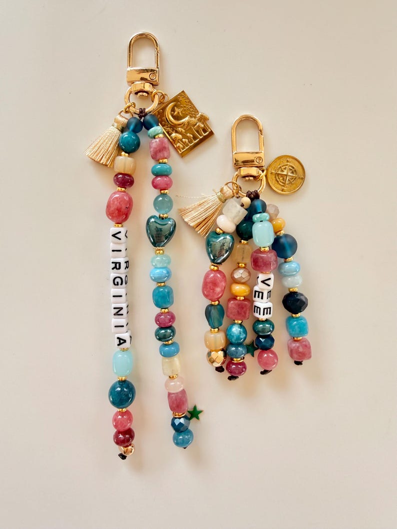 May include: Two beaded keychains with gold-tone hardware. One keychain spells "VIRGINIA" with white letter beads. The other keychain spells "VEE". Both keychains feature colorful beads, tassels, and charms, including a gold star and a crescent moon.