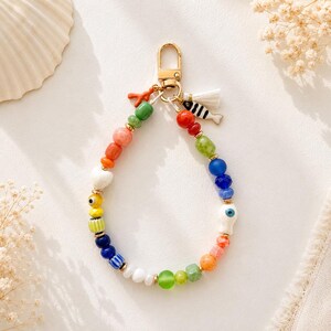 May include: A beaded keychain featuring a gold-tone lobster clasp and a variety of colorful beads in shades of red, orange, yellow, green, blue, and white. The accessory includes small charms shaped like a coral branch, a white tassel, and a fish.