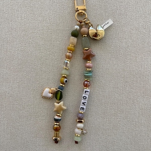 May include: A gold-toned keychain featuring two strands of multi-colored beads, including spherical, star-shaped, and evil eye beads. One strand includes white letter beads spelling LOVE. A gold fortune cookie charm and a white heart charm are attached.
