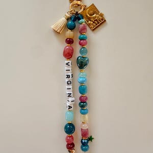 May include: A beaded keychain with a gold clasp, tassel, and charm. The keychain features beads in various colors, including red, blue, and green. The word "VIRGINIA" is spelled out with white letter beads. A green star charm hangs at the bottom.