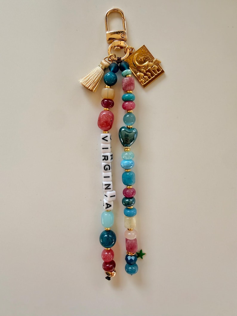 May include: A beaded keychain with a gold clasp, tassel, and charm. The keychain features beads in various colors, including red, blue, and green. The word "VIRGINIA" is spelled out with white letter beads. A green star charm hangs at the bottom.