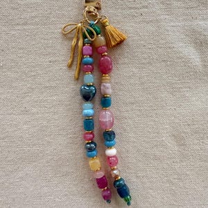 May include: A colorful beaded keychain with a gold-tone clasp, bow, and tassel. The beads are in various shapes and sizes, including heart-shaped and round beads in shades of pink, blue, green, and yellow. The keychain is a decorative accessory.