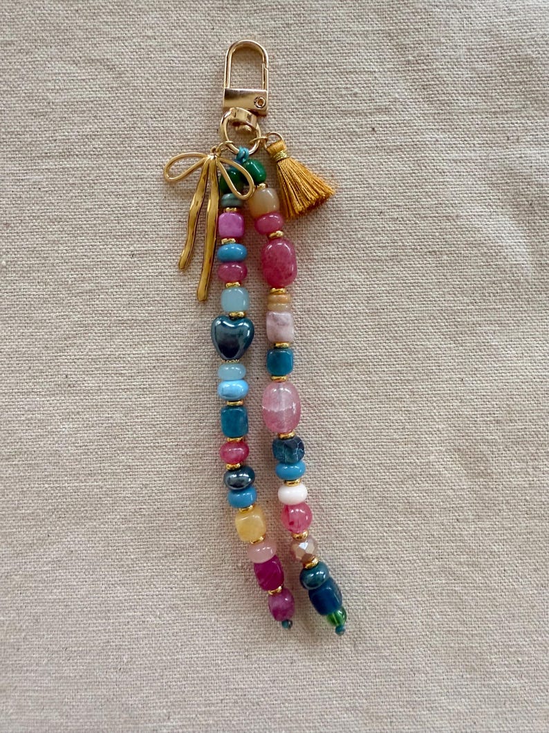 May include: A colorful beaded keychain with a gold-tone clasp, bow, and tassel. The beads are in various shapes and sizes, including heart-shaped and round beads in shades of pink, blue, green, and yellow. The keychain is a decorative accessory.