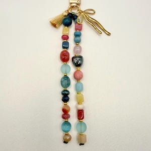 May include: A beaded keychain with a gold-tone clasp and accents. The keychain features strands of colorful beads in various shapes and sizes, including red, blue, yellow, and pink. A small gold tassel and a decorative gold bow add detail.