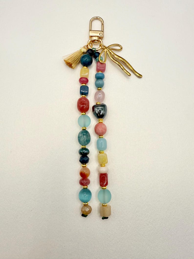 May include: A beaded keychain with a gold-tone clasp and accents. The keychain features strands of colorful beads in various shapes and sizes, including red, blue, yellow, and pink. A small gold tassel and a decorative gold bow add detail.