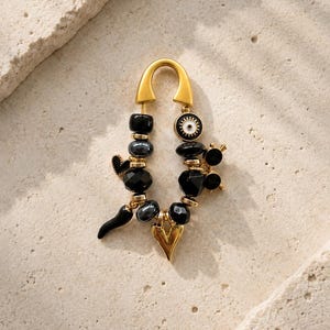 May include: Gold-toned earring with a curved top and dangling charms. The charms include black beads, a heart, a horn, and an eye-shaped design. The earring is set against a neutral, textured background.