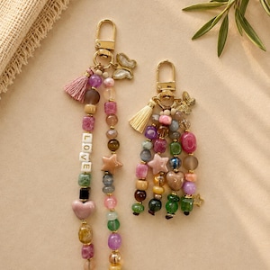 May include: Two gold-toned metal keychains featuring multi-colored beads in various shapes including hearts and stars. The longer keychain includes white letter beads spelling LOVE and a pink tassel. The shorter keychain features a cream tassel.