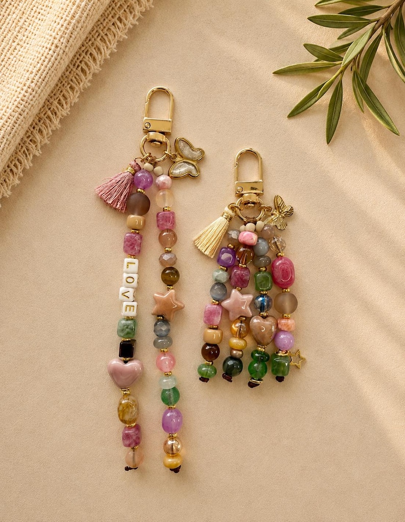 May include: Two gold-toned metal keychains featuring multi-colored beads in various shapes including hearts and stars. The longer keychain includes white letter beads spelling LOVE and a pink tassel. The shorter keychain features a cream tassel.