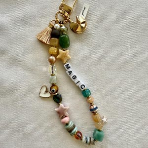 May include: A gold keychain with a charm that says "good luck!" and a beaded charm with the word "MAGIC" spelled out in white letters. The beads are green, brown, white, pink, and gold.