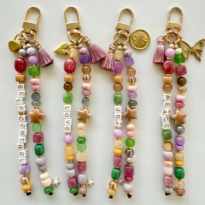 May include: Four beaded keychains with gold-tone lobster clasps. Each features two strands of multi-colored beads, including letter beads spelling SELF CONTROL, LOVE, JOY, and PEACE. Accents include tassels, charms like hearts, stars, and butterflies.