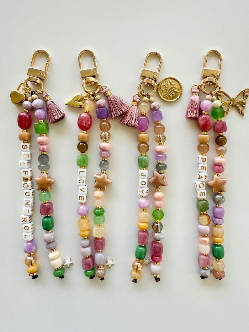 May include: Four beaded keychains with gold-tone lobster clasps. Each features two strands of multi-colored beads, including letter beads spelling SELF CONTROL, LOVE, JOY, and PEACE. Accents include tassels, charms like hearts, stars, and butterflies.