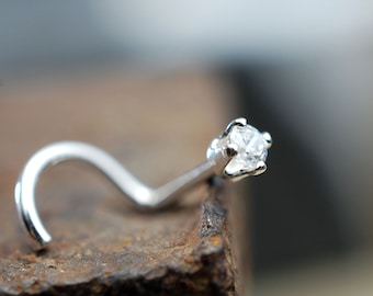 May include: A silver-toned nose stud featuring a single clear, round-cut gemstone held in a four-prong setting. The metal post is curved into a hook shape, designed for secure placement, with a polished finish that reflects light on the stone.
