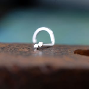 以下が含まれることがあります： 湾曲したポストと、並んで配置された2つの小さな滑らかな円形の球体で構成される装飾的なエンドピースを備えたシルバーのノーズリング。金属は磨かれた反射仕上げで、曲げられたワイヤー構造のデザインになっています。
