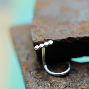 May include: A silver-toned nose ring featuring a curved post and a horizontal bar adorned with four small, evenly spaced circular beads. The jewellery piece has a polished metallic finish and a simple, clean design with a curved end for secure placement.