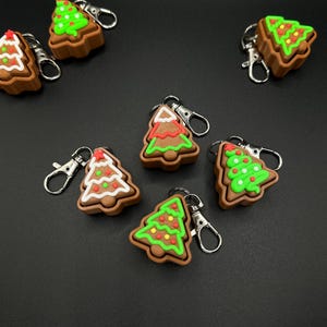 May include: A collection of Christmas tree-shaped keychains. The keychains are brown with green, red, and white detailing, resembling gingerbread cookies. Each keychain has a silver-colored clasp.