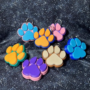 May include: Seven colorful paw print keychains with metal rings. The paw prints are made of a soft, textured material and come in a variety of colors, including blue, orange, purple, and beige.