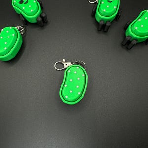 May include: Green pickle-shaped keychains with white polka dots and black legs. Some keychains have sunglasses. The keychains are made of plastic and have a silver clasp. The keychains are on a black surface.