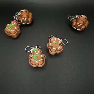May include: Several gingerbread man keychains with silver key rings. The gingerbread men are brown with white icing details, green scarves, and red bows. The keychains are on a black background.