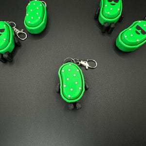 May include: Green pickle-shaped keychains with white polka dots and black arms and legs. Some pickles have black sunglasses. Each keychain has a silver clasp for attaching to keys or bags. The keychains are on a dark gray surface.