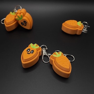 May include: Orange carrot-shaped keychains with green tops and smiling faces. The keychains have silver metal clasps and rings. The background is a solid black color.