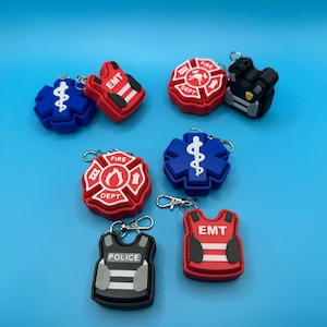 May include: A collection of keychains featuring emergency service symbols. The keychains include designs for EMT, Fire Dept, Police, and medical symbols. The keychains are in red, blue, black, and white colors. Each keychain has a silver-colored keyring.