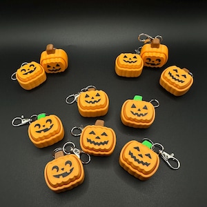 May include: A collection of orange pumpkin-shaped keychains with black carved faces and brown or green stems. Each keychain has a silver clasp. The keychains are arranged on a black surface, perfect for Halloween.