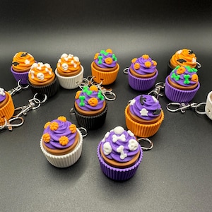 K&ouml;nnte beinhalten: Eine Sammlung von Halloween-Themen-Schl&uuml;sselanh&auml;ngern in Form von Cupcakes. Jeder Cupcake hat ein einzigartiges Design, darunter orangefarbene K&uuml;rbisse, wei&szlig;e Totenk&ouml;pfe und gr&uuml;ne Ranken. Die Cupcakes sind in verschiedenen Farben erh&auml;ltlich, darunter Orange, Lila, Schwarz und Wei&szlig;. Jeder Schl&uuml;sselanh&auml;nger hat einen silbernen Verschluss.