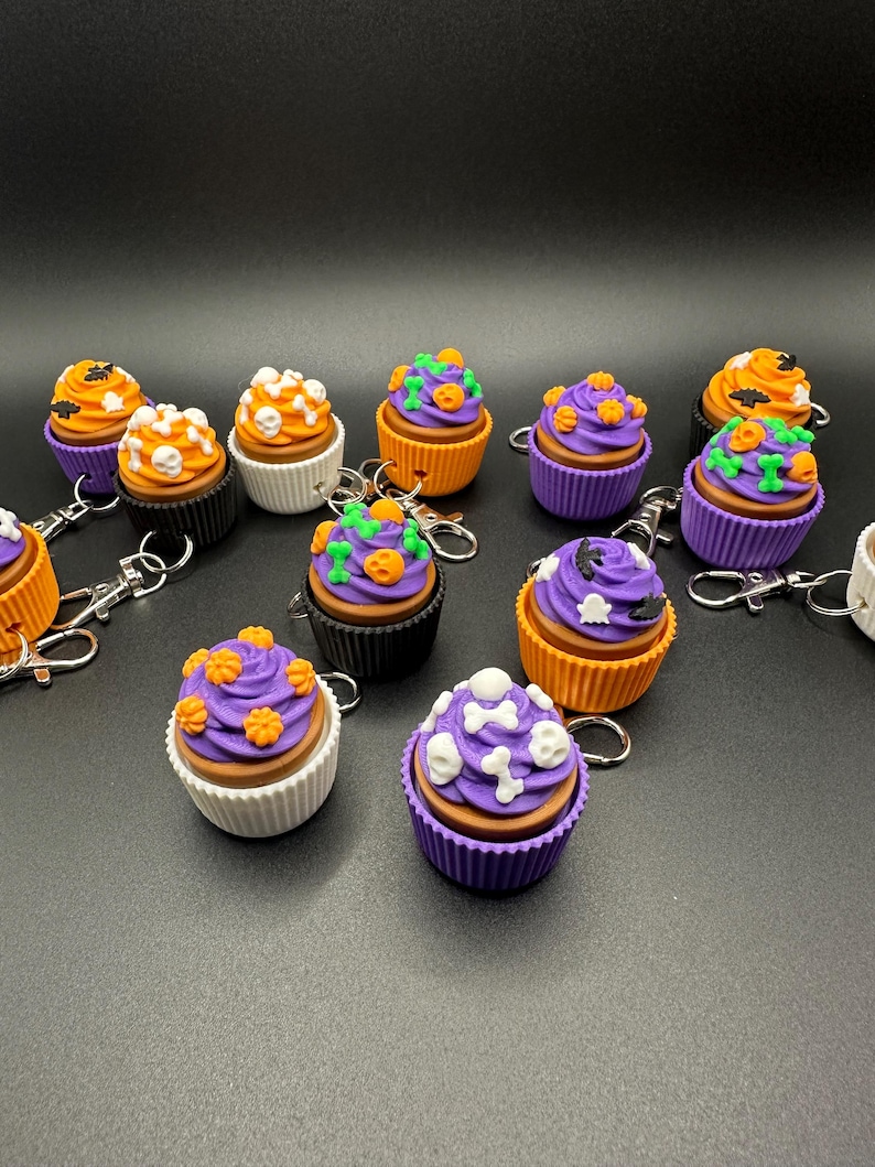 K&ouml;nnte beinhalten: Eine Sammlung von Halloween-Themen-Schl&uuml;sselanh&auml;ngern in Form von Cupcakes. Jeder Cupcake hat ein einzigartiges Design, darunter orangefarbene K&uuml;rbisse, wei&szlig;e Totenk&ouml;pfe und gr&uuml;ne Ranken. Die Cupcakes sind in verschiedenen Farben erh&auml;ltlich, darunter Orange, Lila, Schwarz und Wei&szlig;. Jeder Schl&uuml;sselanh&auml;nger hat einen silbernen Verschluss.